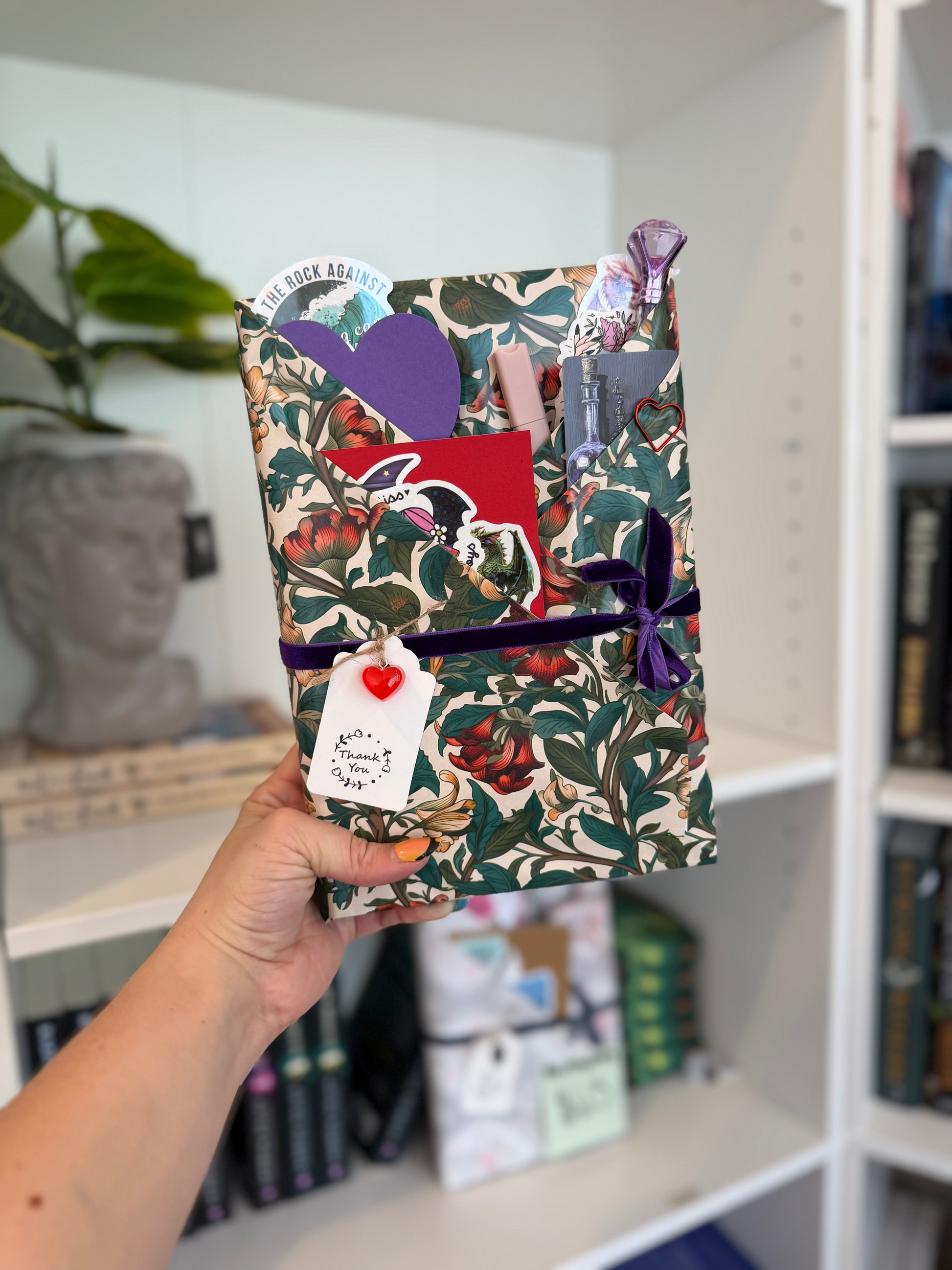 Hand holding a floral-patterned wrapping paper with decorative elements against a bookshelf background
